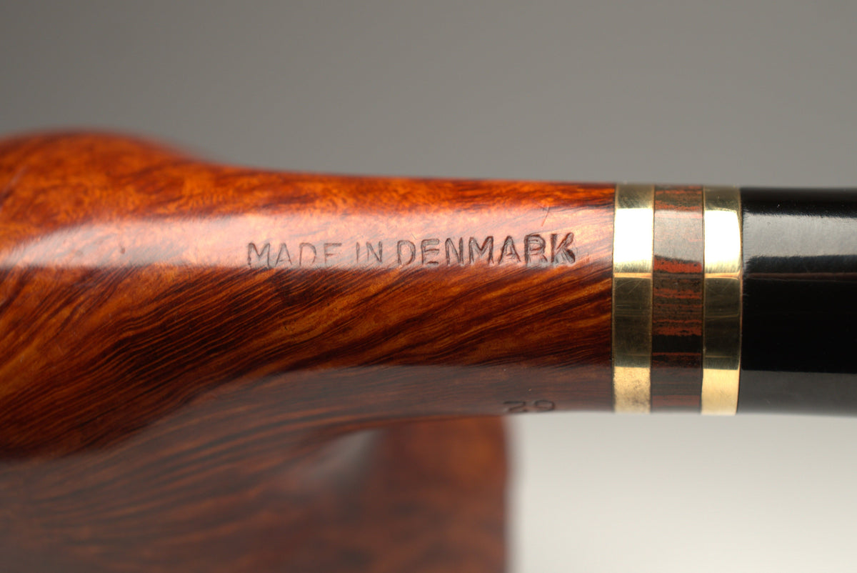 Close-up of a polished wooden smoking pipe handle with a glossy black ferrule and two gold metal bands, featuring 'MADE IN DENMARK' engraved on the wood, set against a neutral gray background.