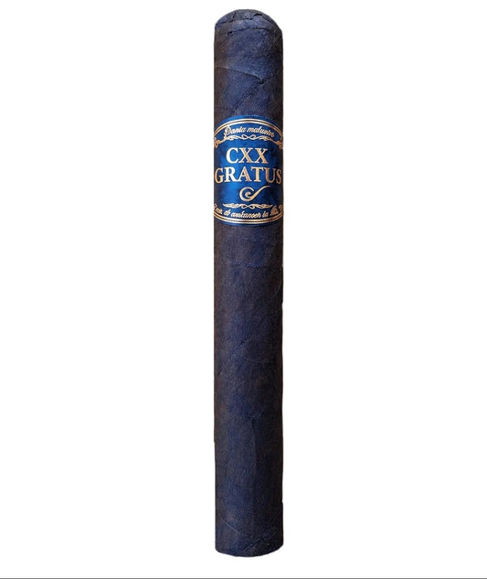 A single dark blue-gray cigar standing upright on a wooden table, with a blue and gold band reading CXX GRATUIT. The background shows a softly blurred cigar room with shelves and rolled cigars.