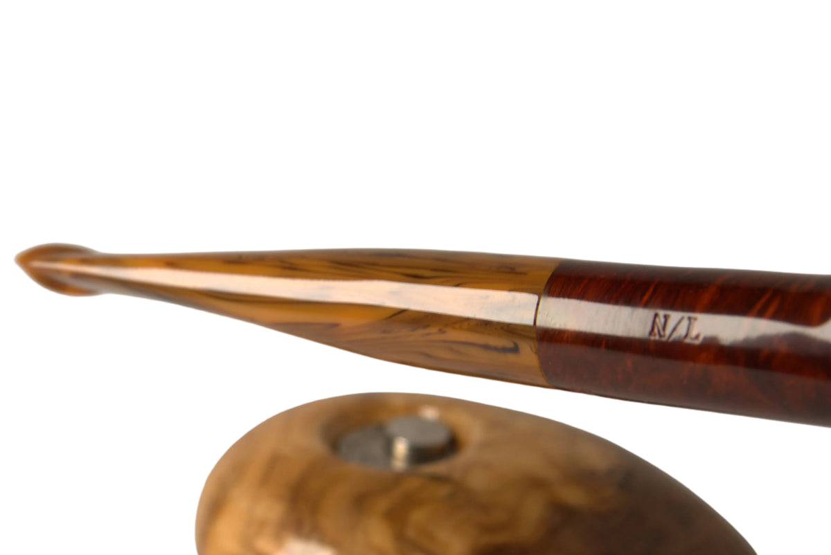A close-up of a polished wooden smoking pipe with a curved stem and a small round wooden stand, held against a neutral gray background. A finger is visible holding the pipe.