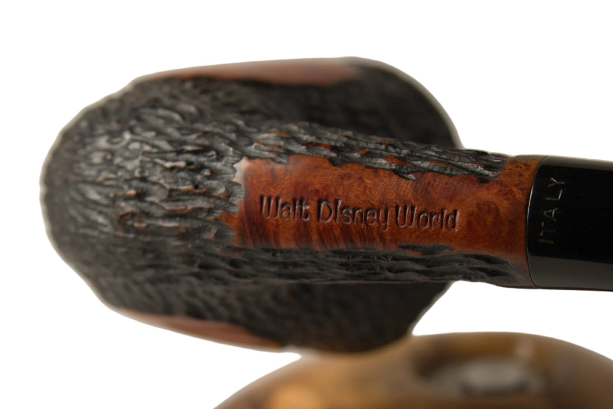 A close-up studio shot of a smoking pipe with a dark ribbed bowl, a black stem, and a brown leather band engraved with 'Walt Disney World'. A polished wooden stand is visible in the blurred background against a neutral light gray gradient.