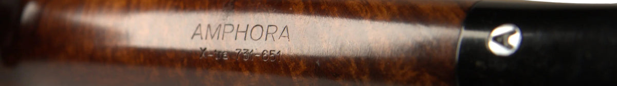 A close-up of a dark brown wooden smoking pipe with 'AMPHORA' engraved on the shank, resting on a tan leather stand against a neutral gray background.