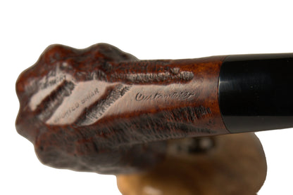 A close-up of a carved smoking pipe bowl with a dark brown wooden stem and a black ferrule, resting on a small round stand against a neutral light gray background.
