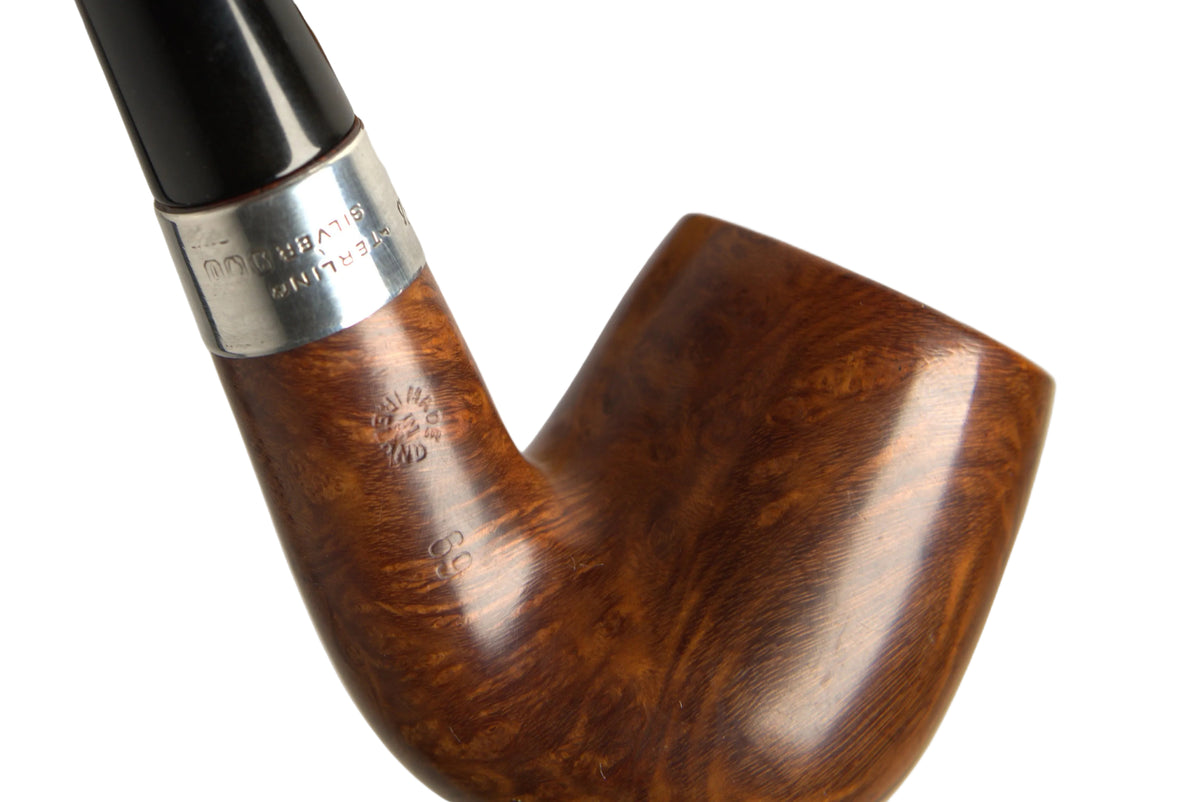 A close-up of a smoking pipe with a polished brown wooden bowl and a black stem, held by a hand against a neutral gray background. A small silver band near the base of the stem has engraved text, and a blurred wooden stand is visible behind the pipe.