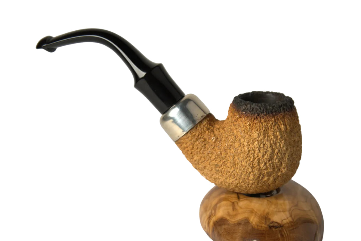 A rustic smoking pipe with a textured light brown wooden bowl, a dark brown blackened tip, a silver metal collar, and a curved black stem, displayed on a carved wooden stand against a neutral gray background.