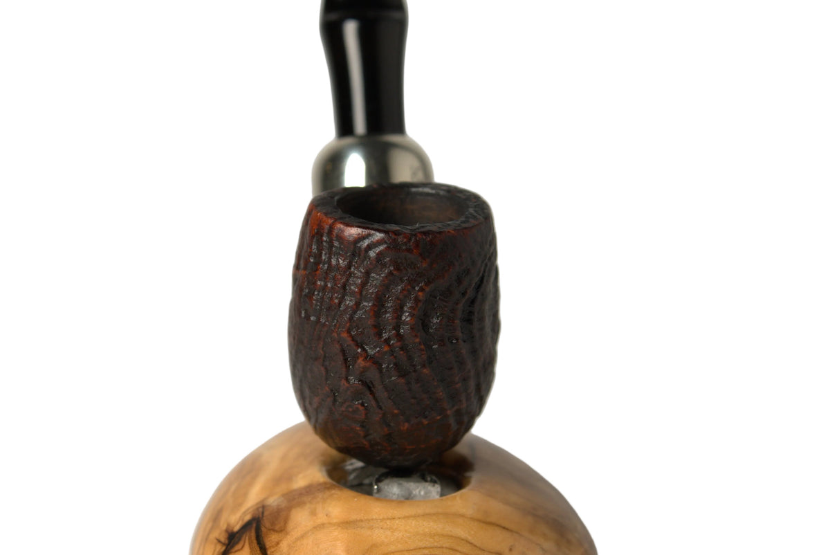 A smoking pipe with a dark brown carved wooden bowl and a black stem, resting on a polished light brown wood base against a neutral gray background.