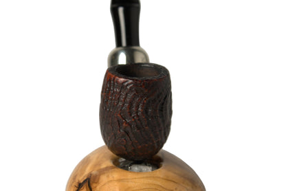 A smoking pipe with a dark brown carved wooden bowl and a black stem, resting on a polished light brown wood base against a neutral gray background.