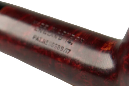 A close-up of a smoking pipe with a glossy dark reddish-brown wooden bowl and a black stem, held by a hand above a small wooden stand on a neutral gray background.