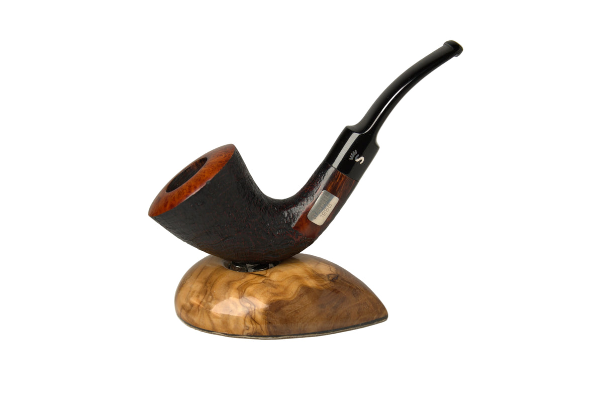 A smoking pipe with a dark brown textured body, reddish-brown rim, and a curved black stem, displayed on a polished burl wood stand against a neutral gray background.