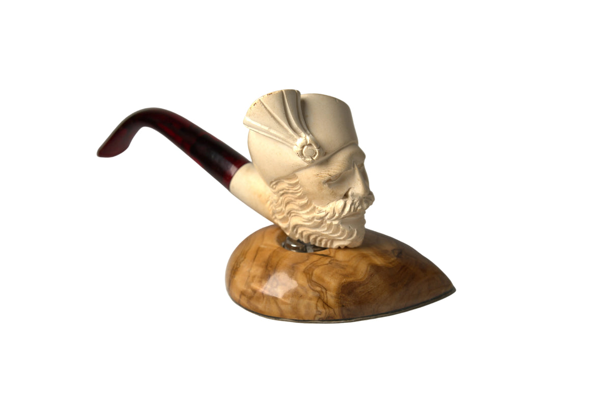 A carved smoking pipe shaped like a bearded man's head wearing a hat, mounted on a polished wood base with a dark reddish-brown stem, photographed on a neutral gray studio background.