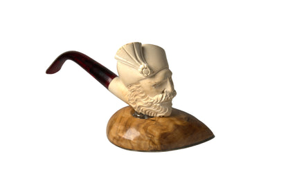 A carved smoking pipe shaped like a bearded man's head wearing a hat, mounted on a polished wood base with a dark reddish-brown stem, photographed on a neutral gray studio background.