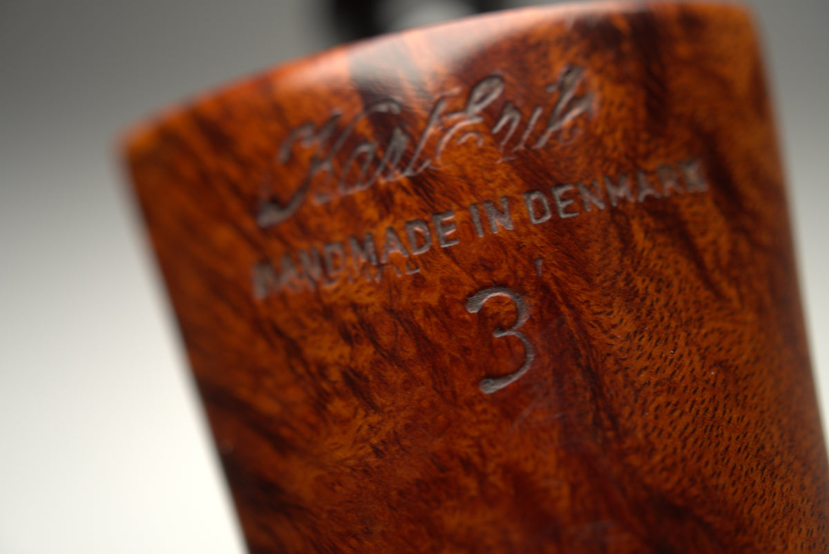 Close-up of a handcrafted wooden smoking pipe body with a richly figured brown wood grain, featuring engraved text that reads 'Kolding HANDMADE IN DENMARK' and a size '3'.