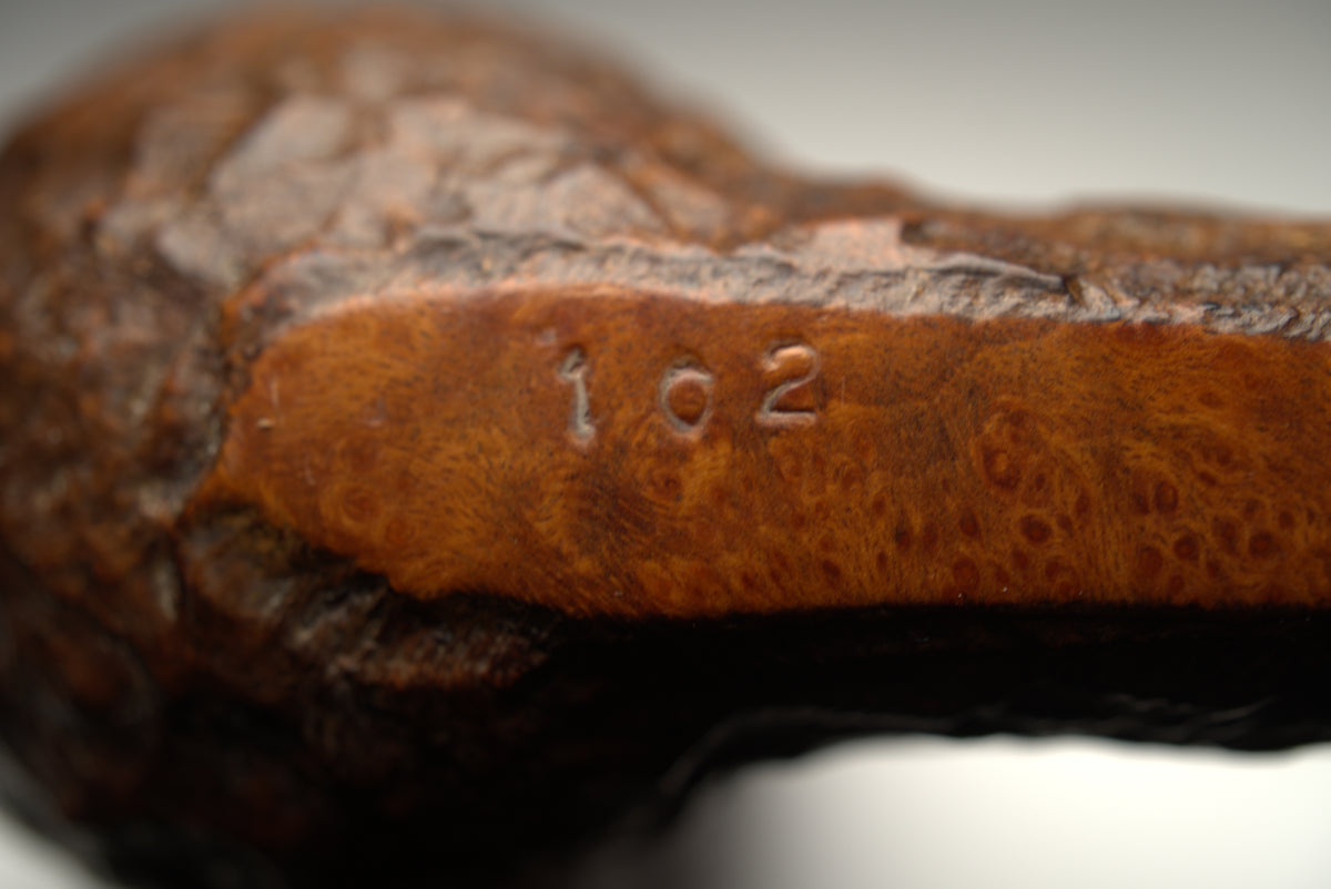 Close-up of a carved wooden object showing a warm brown wood grain surface with the engraved numbers '102' on a soft blurred neutral background.