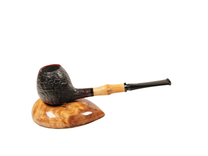 A smoking pipe with a dark textured bowl, a light tan bamboo-style stem, and a black mouthpiece, displayed on a carved wooden stand against a plain white background.