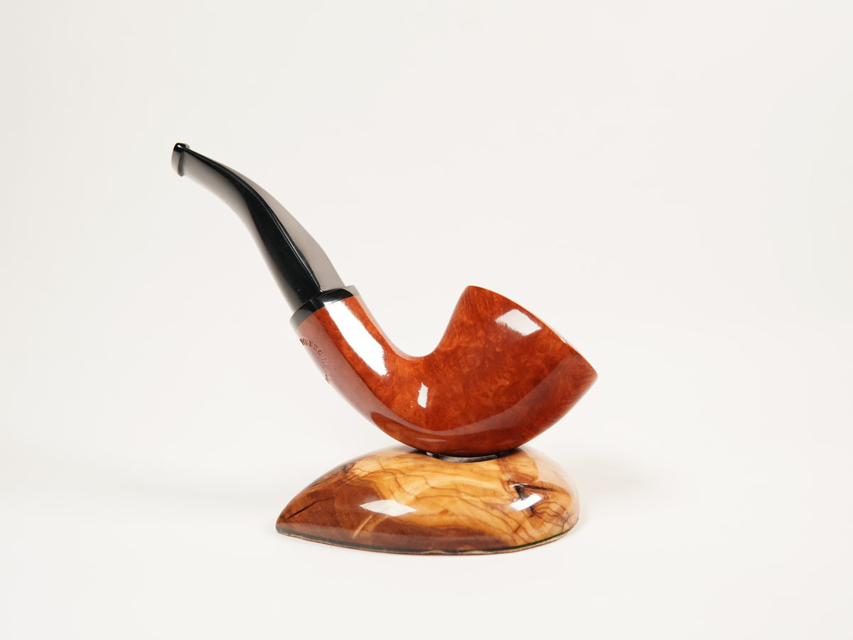 A polished smoking pipe with a curved reddish-brown wooden bowl and a black stem, displayed on a matching carved wooden stand against a plain white background.