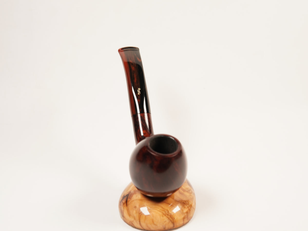 A smoking pipe with a dark reddish-brown glossy stem and a round dark brown bowl, resting on a light tan wood base against a plain white background.