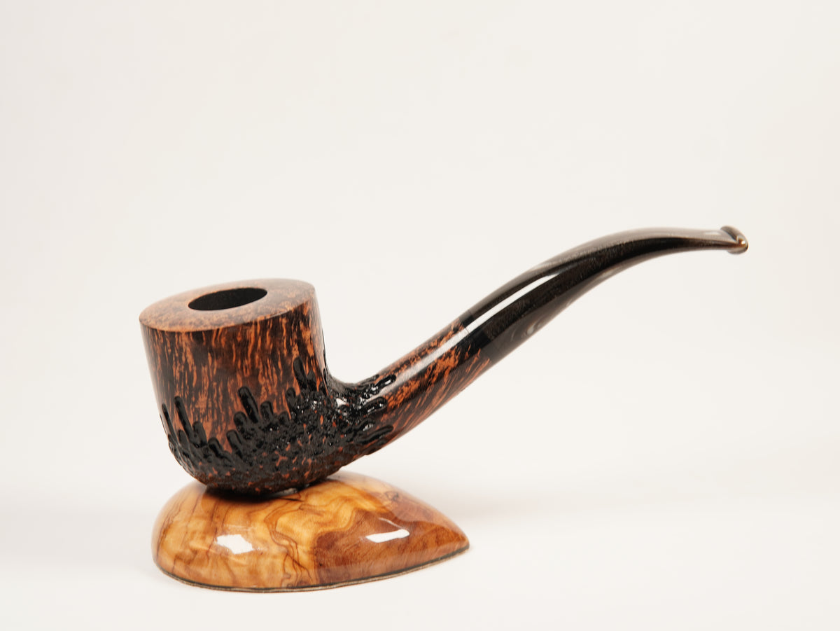 A smoking pipe with a dark brown and black carved stem and bowl, resting on a polished burl wood display stand against a plain white background.