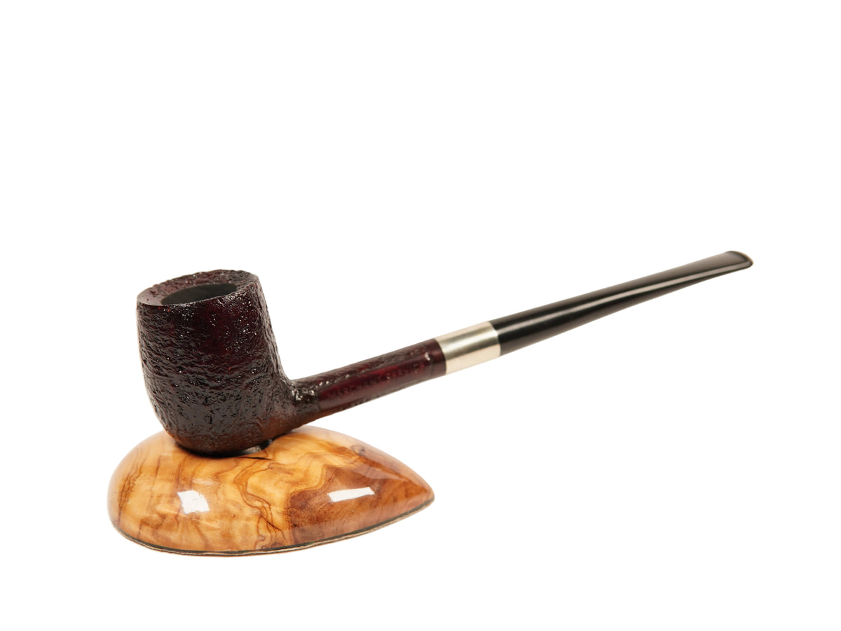 A dark brown smoking pipe with a textured bowl and a long black stem, resting on a polished wood stand with a swirling tan and brown grain pattern, set against a plain white background.