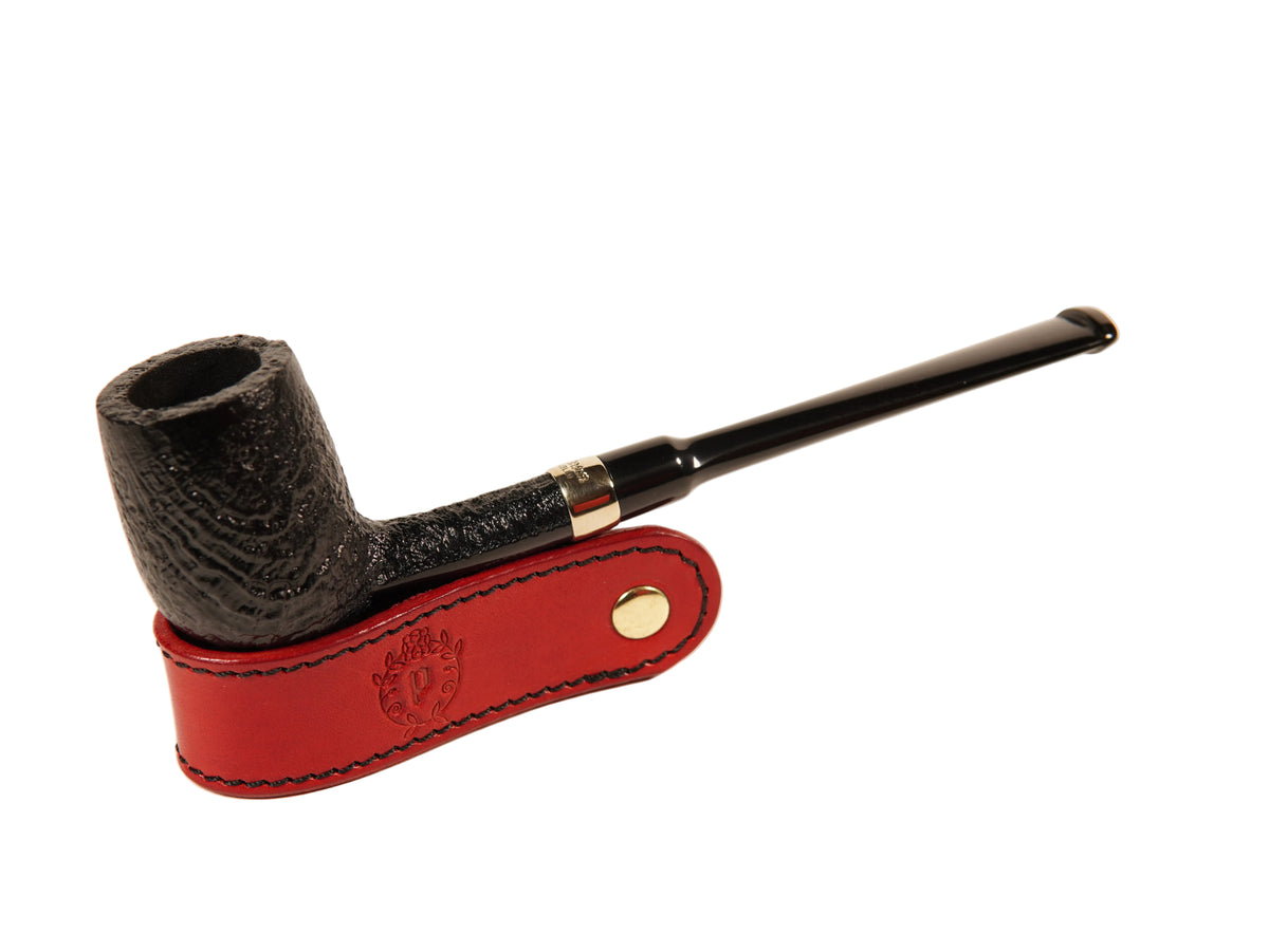 A dark textured smoking pipe with a long black stem, resting on a red leather holder with gold-tone hardware, photographed on a clean off-white background.