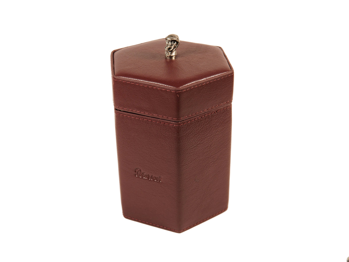 A dark reddish-brown textured leather hexagonal box with a matching lid and a small silver skull finial on top, featuring an embossed logo on the front, set against a plain light background.