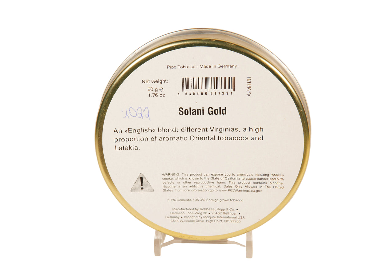 A round tin of Ammu Solani Gold pipe tobacco on a clear display stand against a warm off-white background. The white label features a barcode, product details, and a small warning icon, framed by a metallic gold rim.