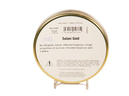 A round tin of Ammu Solani Gold pipe tobacco on a clear display stand against a warm off-white background. The white label features a barcode, product details, and a small warning icon, framed by a metallic gold rim.