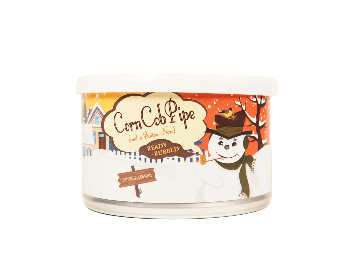 A white plastic tub of Corn Cob Pipe ready-rubbed balm with a colorful winter-themed label featuring a snowman, house, and autumn tree on a white background.
